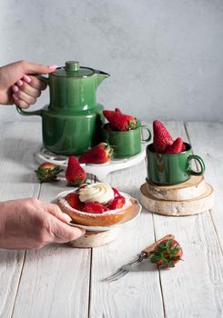 Fruit still life with strawberries and green tea set, strawberry cake with cream sweet dessert, female hand breaks off a piece of cake with a dessert fork. High quality photo