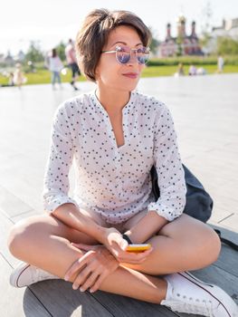 Woman in colorful sunglasses sits on wooden open stage in public park. Pretty female in casual clothes texting on smartphone.