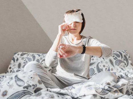 Sleepy woman in grey pajama and sleeping mask on face. She is just woke up and sit in bed. Early morning in cozy home.