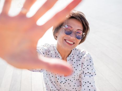 Wind ruffles short hair of freckled woman in colorful sunglasses. Smiling woman at open wooden scene of urban park. Summer vibes. Sincere emotions.