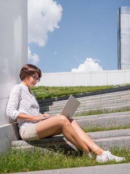 Woman sits with laptop on urban park bench. Freelancer at work. Student learns remotely from outdoors. Modern lifestyle. Summer vibes. Outdoor workplace.