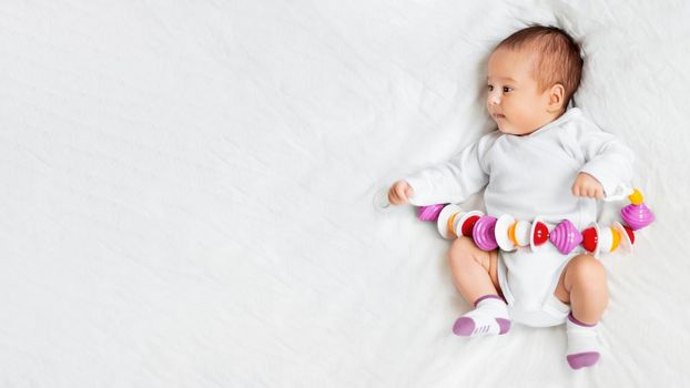 Baby girl holds colorful rattle toy. Top view of little kid lying on crumpled white linen with his first toy. Newborn in bed. Banner with copy space.