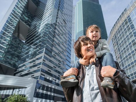 Little boy sits on father's shoulders between skyscrapers. Dad and son looks on glass walls of buildings. Future and modern technologies, life balance and family life in well keeps districts.