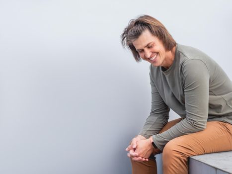 Man with long hair and happy look sits near grey wall. Smiling man in earth toned casual clothes is laughing on horizontal banner with copy space.