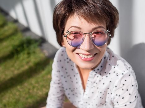 Smiling woman in colorful sunglasses has a rest on lawn in urban park. Nature in town. Relax outdoors after work. Summer vibes.