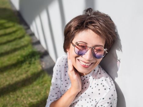 Smiling woman in colorful sunglasses has a rest on lawn in urban park. Nature in town. Relax outdoors after work. Summer vibes.