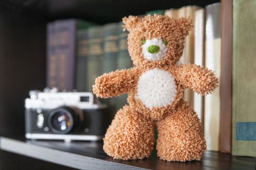 Hand made teddy bear on bookshelf. Crocheted brown toy for children. Cute souvenir, handicraft product.