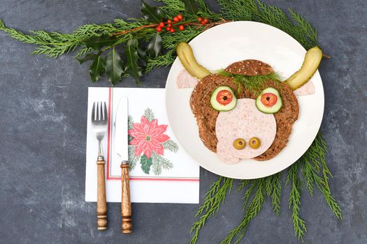 Sandwich in the form of a portrait of a cheerful bull made of dark bread, sausages and vegetables on a plate, on a dark background with a decor of thuja and holly branches