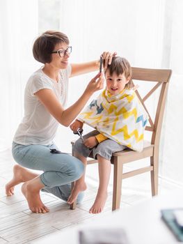 Mother cuts her son's hair by herself. Little boy sits, covered with cloth, and holds pair of scissors. Family life.