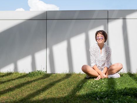 Smiling woman in colorful sunglasses has a rest on lawn in urban park. Nature in town. Relax outdoors after work. Summer vibes.