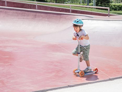 Little boy rides kick scooter in skate park. Special concrete bowl structures in urban park. Training to skate at summer.