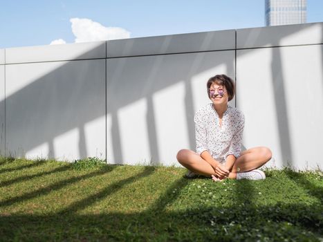 Smiling woman in colorful sunglasses has a rest on lawn in urban park. Nature in town. Relax outdoors after work. Summer vibes.