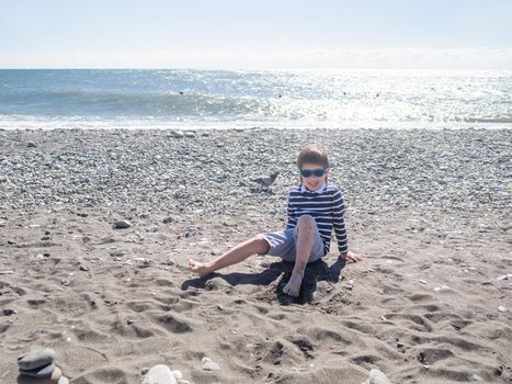 Happy boy is playing with wet sand, stones and driftwood on pebble beach. Vacation on seaside. Exploring nature in childhood.