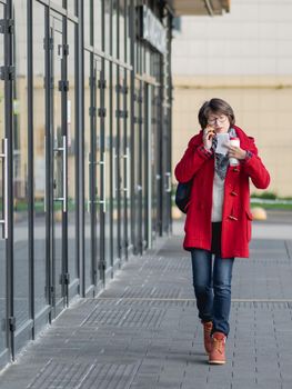 Woman in red duffle coat talks by smartphone. Woman walks down the street grasping cup of take away coffee and paper cheque.