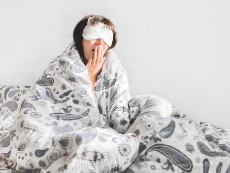 Woman in grey pajama and sleeping mask on face. She is just woke up and sit in bed. Early morning in cozy home.