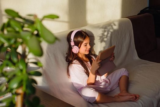 A little adorable girl in pink home clothes and pink headphones sits on the couch, looks into a digital tablet.