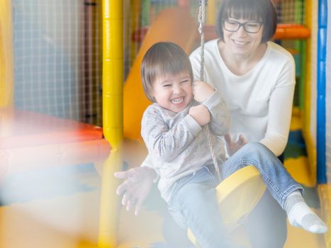 Toddler plays on rope swing with his mother or babysitter. Physical development for little children. Interior of kindergarten or nursery.