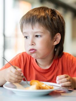 Fussy kid in red t-shirt eats pancakes with knife and fork. Little boy with funny expression on face. Tasty pastry for breakfast.