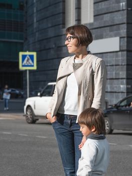 Mother and son stand on street in sunlight. Woman and kid in modern town with transport traffic and road sign. Family in city.