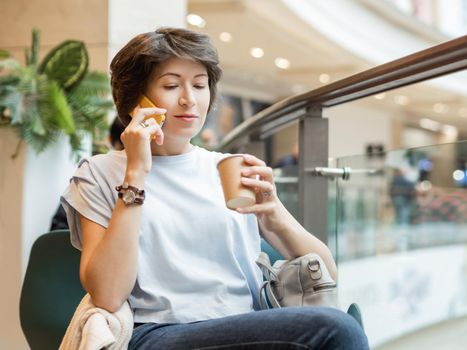 Woman is talking by smartphone and drinking coffee at shopping mall. Coffee break at store.