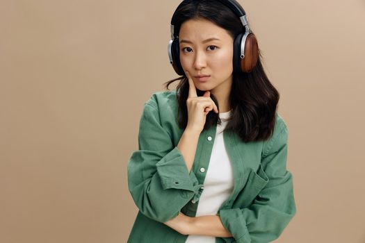 Pensive thoughtful cute Asian student young woman in khaki green shirt headphones recline on hand posing isolated on beige pastel studio background. Cool fashion offer. Music App Platform Ad concept