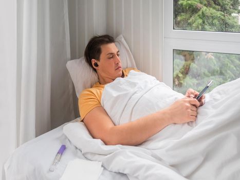 Sick man is lying in bed with thermometer. Man is searching online for disease symptoms or texting to his doctor. Using wireless technology in case of temporary disability.