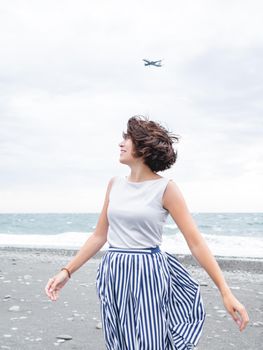 Plane flies in cloudy sky over smiling woman on seaside. Woman with hair ruffled with the wind. Wanderlust concept. Vacation on sea coast.