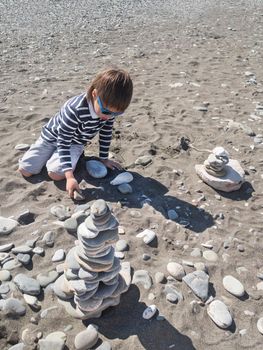 Little boy is building pyramid with stones and driftwood on pebble beach. Vacation on seaside. Exploring nature in childhood.