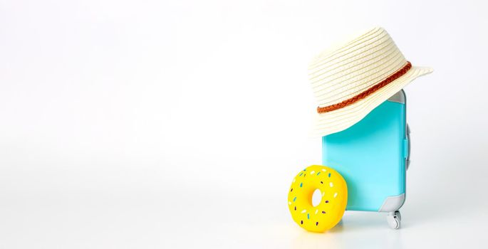 A blue suitcase on wheels, a straw hat, and a yellow inflatable donut stand on a white background. copy space. Wide banner