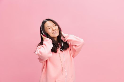 Relaxing Asian young lady in pink hoodie sweatshirt with cute headphones listen fav song closing eyes posing isolated on over pink studio background. Good offer. Sound streaming platform ad concept