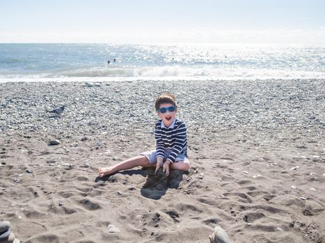 Happy boy is building pyramid with wet sand, stones and driftwood on pebble beach. Vacation on seaside. Exploring nature in childhood.