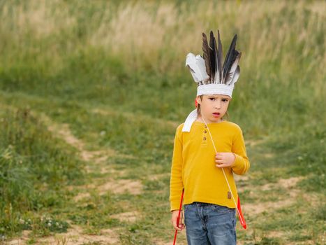 Portrait of boy playing American Indian on field. Kid has handmade headdress made of feathers and bow with arrows. Costume role play. Outdoor leisure activity.