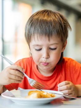 Little kid in red t-shirt eats pancakes with knife and fork. Curious boy with puzzled expression on face. Tasty pastry for breakfast.