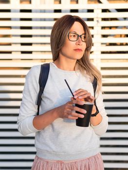 Woman with cup of coffee meets sunset on wooden pier. Female with curly hair and eyeglasses enjoys nature outdoors. Summer casual clothes. Summer vibes.