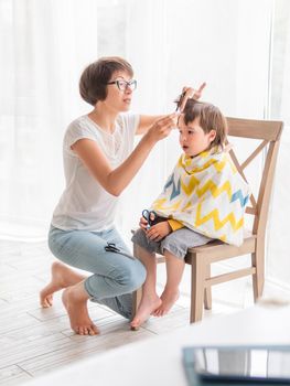 Mother cuts her son's hair by herself. Little boy sits, covered with cloth, and holds pair of scissors. New normal in case of coronavirus COVID-19 quarantine. Beauty at home lifestyle.