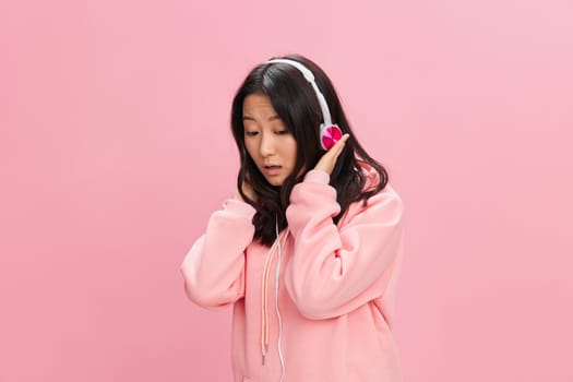 Upset confused Asian student young lady in pink hoodie sweatshirt with cute headphones looks at you posing isolated on over pink studio background. Good offer. Sound streaming platform ad concept