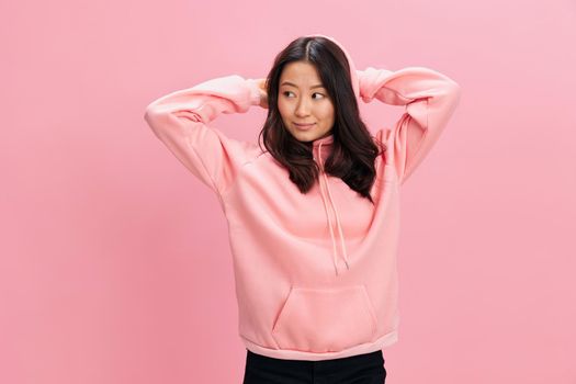 Smiling happy cute Asian student young lady in pink hoodie sweatshirt hold hands behind head posing isolated on over pink studio background. Good offer. Fashion New Collection concept