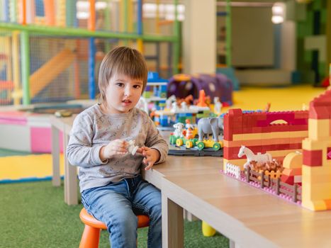 Toddler plays with colorful toy blocks. Little boy with figure from toy constructor. Interior of kindergarten or nursery. Indoors leisure activity for children.