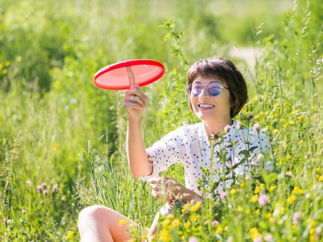 Woman in colorful sunglasses plays with red frisbee, enjoys sunlight and flower fragrance on grass field. Summer vibes. Relax outdoors. Self-soothing.