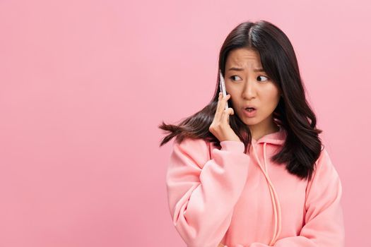Irritated angry cute Asian student young lady in pink hoodie sweatshirt quarrel with boyfriend on phone posing isolated on pink studio background. Good offer. Gadget addiction Social Media concept