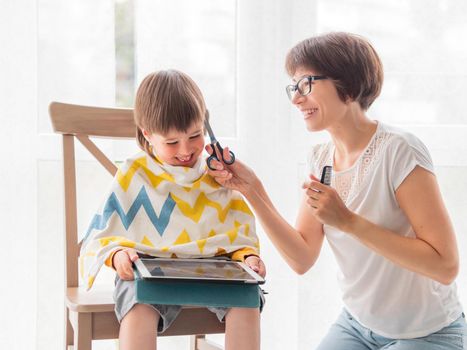 Mother cuts her son's hair by herself. Little boy sits, covered with cloth, and holds digital tablet. New normal in case of coronavirus COVID-19 quarantine and lockdown.