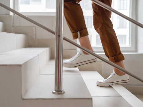 Woman in white sneakers and khaki trousers goes upstairs to her apartment. White staircase in apartment building. Casual outfit, urban fashion.