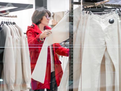 Woman chooses trousers at clothing store. Casual trousers hanging on hangers. View through transparent shop window. Shopping at mall. Basic clothes for everyday wear. Modern urban fashion.