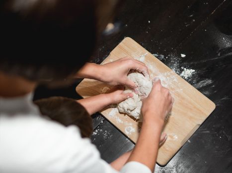 Mother and son are cooking together. Mom and toddler knead dough from flour and eggs on black wooden table. Family time. Fun at kitchen. Developing fine motor skills.