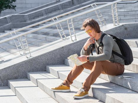 Man sits on stone staircase and reads map. Solo-travelling around city. Urban tourism. Modern architectural landmarks.