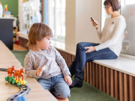 Toddler plays with colorful toy blocks while his mother or babysitter texting in smartphone. Little boy with toy constructor. Interior of kindergarten or nursery.