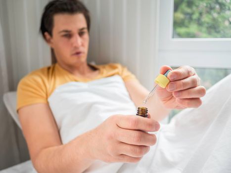 Ill man is lying in bed with liquid medicine. Man is measuring drops. Feeling sick because of disease and temporary disability.