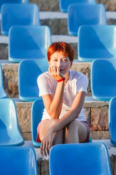 Young woman with short red hair sits relaxed on plastic blue seat in deserted open air audience. Summer vibes.