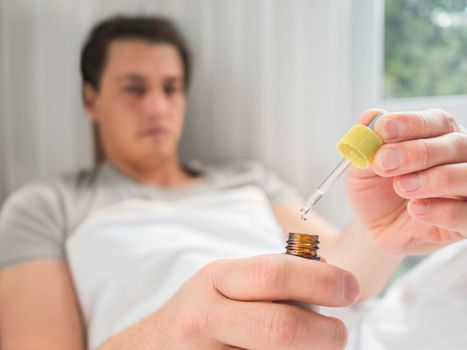 Ill man is lying in bed with liquid medicine. Man is measuring drops. Feeling sick because of disease and temporary disability.