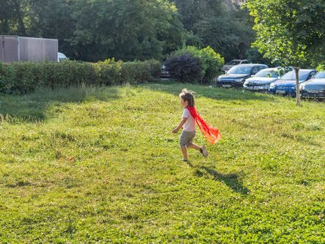 Little boy is playing superhero on lawn. Kid in handmade bright red cloak. Outdoor role-playing game. Costume play at sunlight.